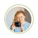 Female photographer with blonde curly hair holding black camera, smiling warmly in light blue sweater